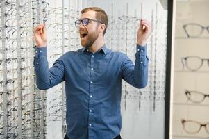 tevreden klant. visie van gelukkig jong mannetje cliënt vervelend nieuw bril, staand in de buurt rek en vitrine met brillen. glimlachen Mens proberen Aan bril foto