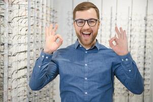 in optiek winkel. portret van mannetje cliënt Holding en vervelend verschillend bril, kiezen en proberen Aan nieuw bril Bij optisch op te slaan. Mens plukken kader voor visie correctie, detailopname. foto