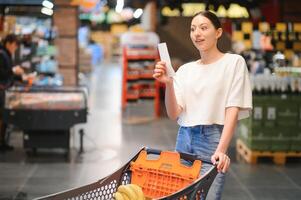 vrouw Bij supermarkt Holding een vol boodschappen doen kar en een boodschappen doen lijst foto