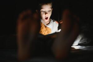 kind lezing boek in bed. kinderen lezen Bij nacht. weinig jongen met fee verhaal boeken in slaapkamer . onderwijs voor jong kinderen. bedtijd verhaal in de avond. schattig kind onder deken in donker kamer met lamp foto