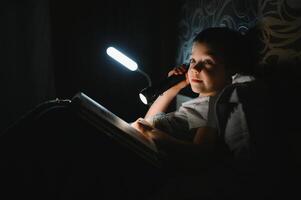 portret van schattig weinig jongen lezing in bed met zaklamp in donker kamer, genieten van sprookjes. foto