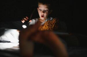 portret van schattig weinig jongen lezing in bed met zaklamp in donker kamer, genieten van sprookjes. foto