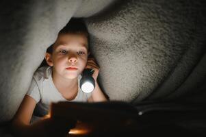 kind lezing boek in bed. kinderen lezen Bij nacht. weinig jongen met fee verhaal boeken in slaapkamer . onderwijs voor jong kinderen. bedtijd verhaal in de avond. schattig kind onder deken in donker kamer met lamp foto