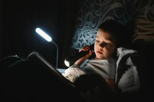 lezing boek en gebruik makend van zaklamp. jong jongen in gewoontjes kleren aan het liegen naar beneden in de buurt tent Bij avond tijd foto