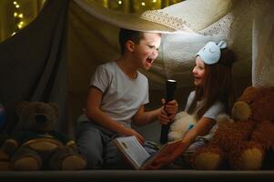 kinderen jongen en meisje spelen en schrikken elk andere met zaklamp in tent Bij nacht. foto