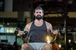 Kaukasisch baard gespierd sport Mens is praktijk gewicht opleiding Aan dubbele halters voor biceps en triceps spier binnen Sportschool met donker achtergrond voor oefenen en training foto