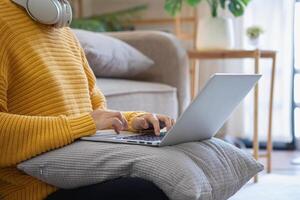 mooi jong vrouw in een geel gewoontjes jurk genieten van luisteren naar muziek- en glimlachen terwijl ontspannende Aan de sofa Bij huis. jong vrouw met hoofdtelefoons toepassingen laptop en smartphone Bij huis. kom tot rust concept foto