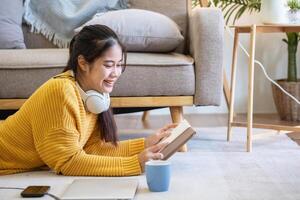 mooi jong vrouw in een geel gewoontjes jurk genieten van luisteren naar muziek- en glimlachen terwijl ontspannende Aan de sofa Bij huis. jong vrouw met hoofdtelefoons toepassingen laptop en smartphone Bij huis. kom tot rust concept foto