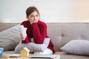 een jong Aziatisch vrouw zit Aan een sofa in haar huis, gevoel bezorgd en gefrustreerd over haar maandelijks uitgaven. divers nut rekeningen. foto