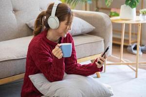 mooi jong vrouw in een rood gewoontjes jurk genieten van luisteren naar muziek- en glimlachen terwijl ontspannende Aan de sofa Bij huis. jong vrouw met hoofdtelefoons toepassingen laptop en smartphone Bij huis. kom tot rust concept. foto