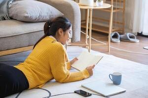 mooi jong vrouw in een geel gewoontjes jurk genieten van luisteren naar muziek- en glimlachen terwijl ontspannende Aan de sofa Bij huis. jong vrouw met hoofdtelefoons toepassingen laptop en smartphone Bij huis. kom tot rust concept foto