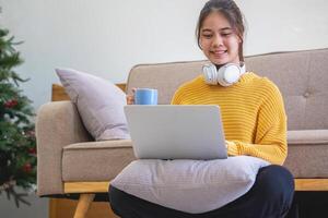 mooi jong vrouw in een geel gewoontjes jurk genieten van luisteren naar muziek- en glimlachen terwijl ontspannende Aan de sofa Bij huis. jong vrouw met hoofdtelefoons toepassingen laptop en smartphone Bij huis. kom tot rust concept foto