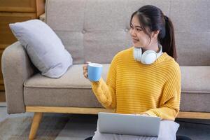 mooi jong vrouw in een geel gewoontjes jurk genieten van luisteren naar muziek- en glimlachen terwijl ontspannende Aan de sofa Bij huis. jong vrouw met hoofdtelefoons toepassingen laptop en smartphone Bij huis. kom tot rust concept foto