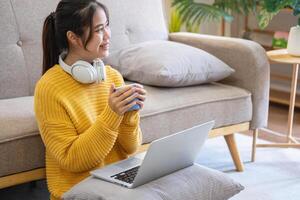 mooi jong vrouw in een geel gewoontjes jurk genieten van luisteren naar muziek- en glimlachen terwijl ontspannende Aan de sofa Bij huis. jong vrouw met hoofdtelefoons toepassingen laptop en smartphone Bij huis. kom tot rust concept foto