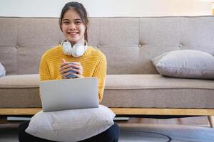 mooi jong vrouw in een geel gewoontjes jurk genieten van luisteren naar muziek- en glimlachen terwijl ontspannende Aan de sofa Bij huis. jong vrouw met hoofdtelefoons toepassingen laptop en smartphone Bij huis. kom tot rust concept foto