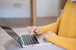 mooi jong vrouw in een geel gewoontjes jurk genieten van luisteren naar muziek- en glimlachen terwijl ontspannende Aan de sofa Bij huis. jong vrouw met hoofdtelefoons toepassingen laptop en smartphone Bij huis. kom tot rust concept foto