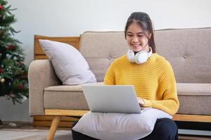 mooi jong vrouw in een geel gewoontjes jurk genieten van luisteren naar muziek- en glimlachen terwijl ontspannende Aan de sofa Bij huis. jong vrouw met hoofdtelefoons toepassingen laptop en smartphone Bij huis. kom tot rust concept foto