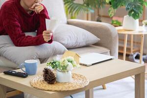 een jong Aziatisch vrouw zit Aan een sofa in haar huis, gevoel bezorgd en gefrustreerd over haar maandelijks uitgaven. divers nut rekeningen. foto