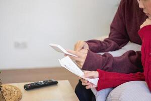 een jong Aziatisch vrouw zit Aan een sofa in haar huis, gevoel bezorgd en gefrustreerd over haar maandelijks uitgaven. divers nut rekeningen. foto