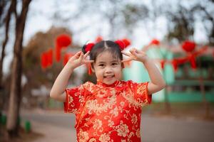 een speels jong meisje in een traditioneel rood cheongsam maken vrede tekens in de buurt haar ogen, met feestelijk decoraties in de achtergrond. foto