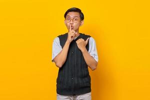 knappe aziatische man die vinger op de lippen maakt, stil gebaar en vinger wijzend naar lege ruimte over gele achtergrond foto