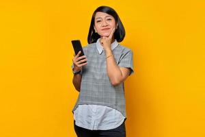 portret van een glimlachende jonge aziatische vrouw die nadenkt over een vraag met de vinger op de kin en een mobiele telefoon vasthoudt foto