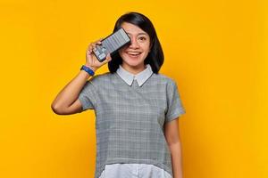 portret van mooie glimlachende aziatische vrouw die een half gezicht bedekt met smartphone geïsoleerd op gele achtergrond foto