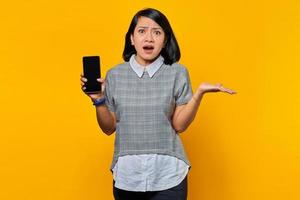 portret van een geschokte jonge aziatische vrouw die een leeg scherm van een smartphone toont en wijst naar een lege ruimte met handpalmen over een gele achtergrond foto