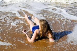 meisje in een blauw het baden pak leugens Aan de kust en ontspant. golven strelen de meisjes lichaam foto