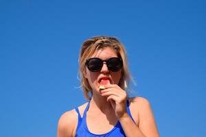 een vrouw is Holding een stuk van watermeloen in de buurt haar mond tegen de blauw lucht. een rijp watermeloen in de handen van een blond foto