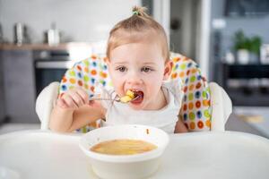 schattig baby meisje kleuter zittend in de hoog stoel en aan het eten haar lunch soep Bij huis keuken. foto