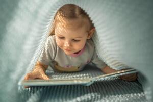 weinig meisje genieten van van recensie een boek onder blauw gebreid plaid in zonnig ochtend- foto