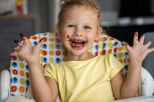 weinig meisje met blond haar- aan het eten eigengemaakt chocola en tonen mond en vuil handen met vlekken van chocola in huis keuken foto