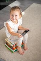 weinig meisje zit Aan stack van kinderen sprookje boeken met smartphone in haar handen foto
