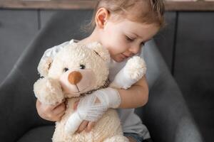 weinig meisje met gebroken vinger houdt teddy beer met een verbonden poot Bij de dokter afspraak in de ziekenhuis foto