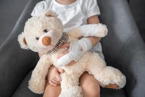 weinig meisje met gebroken vinger houdt teddy beer met een verbonden poot Bij de dokter afspraak in de ziekenhuis foto