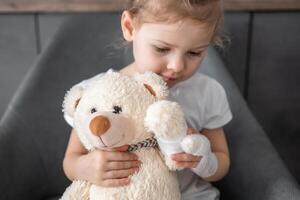 weinig meisje met gebroken vinger houdt teddy beer met een verbonden poot Bij de dokter afspraak in de ziekenhuis foto