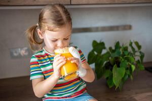 weinig meisje drinken vers sap zittend Aan tafel in huis keuken foto