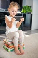 schattig weinig meisje zit Aan een stack van kinderen sprookje boeken en horloges tekenfilms Aan haar smartphone foto