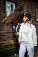 mooi blond professioneel vrouw jockey in wit kleren staand in de buurt bruin paard. vriendschap met paard. hoog kwaliteit foto