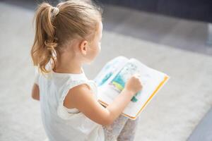 schattig weinig meisje is zittend en bladeren door een boek met afbeeldingen van fee verhalen foto