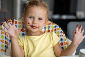 weinig meisje met blond haar- aan het eten eigengemaakt chocola en tonen mond en vuil handen met vlekken van chocola in huis keuken foto