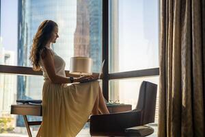 elegant zakenvrouw freelancer werken met laptop, ze is zittend Aan de tafel, genieten van panoramisch visie in venster met stad achtergrond. laag sleutel foto. hoog kwaliteit foto