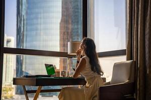 elegant zakenvrouw freelancer met laptop zittend Aan de stoel en genieten van panoramisch visie in de stad achtergrond. laag sleutel foto