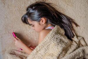 schattig weinig meisje gebruik makend van een smartphone. kind spelen met mobiel telefoon, aan het liegen Aan een bed. vrije tijd. technologie en internet concept. kleuter houdende Aan bed en op zoek Bij smartphone. foto