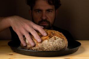 aantrekkelijk jong Kaukasisch chef poseren met wit zuurdesem brood. de zuurdesem brood is de centraal hoofdrolspeler van de tafereel, staand uit met mooi gouden tonen tegen de donker achtergrond. foto