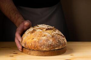 de handen van een jong Mens behandeling zuurdesem brood, markeren de brood met mooi gouden tonen tegen de donker achtergrond. foto