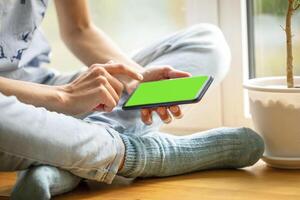 jong meisje in blauw jeans zittend Aan venster doden met smartphone met groen scherm klikken Aan scherm foto