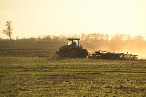 trekker Bij zonsondergang ploeg ploeg een veld. bewerken de bodem in de vallen na oogst. de einde van de seizoen foto