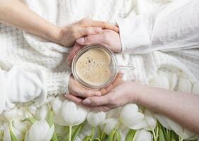 vier handen houden een van hartvormig koffie beker, wit tulpen foto